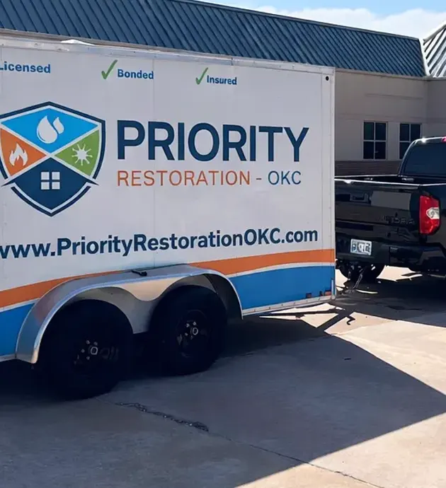 A blue and white enclosed trailer belonging to Priority Restoration OKC is parked in a commercial lot. The trailer displays the company's logo featuring a house shield emblem and website www.PriorityRestorationOKC.com. The vehicle is licensed, bonded, and insured, as indicated by text at the top of the trailer.