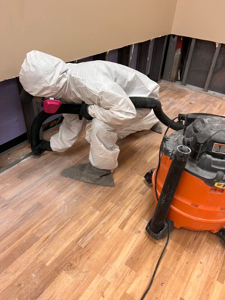 A worker in full protective white coveralls and breathing apparatus bends over wooden flooring while operating an orange and black industrial wet/dry vacuum. The workspace appears to be an interior renovation or remediation project, with plastic sheeting visible on the walls and various equipment scattered across the wood floor.