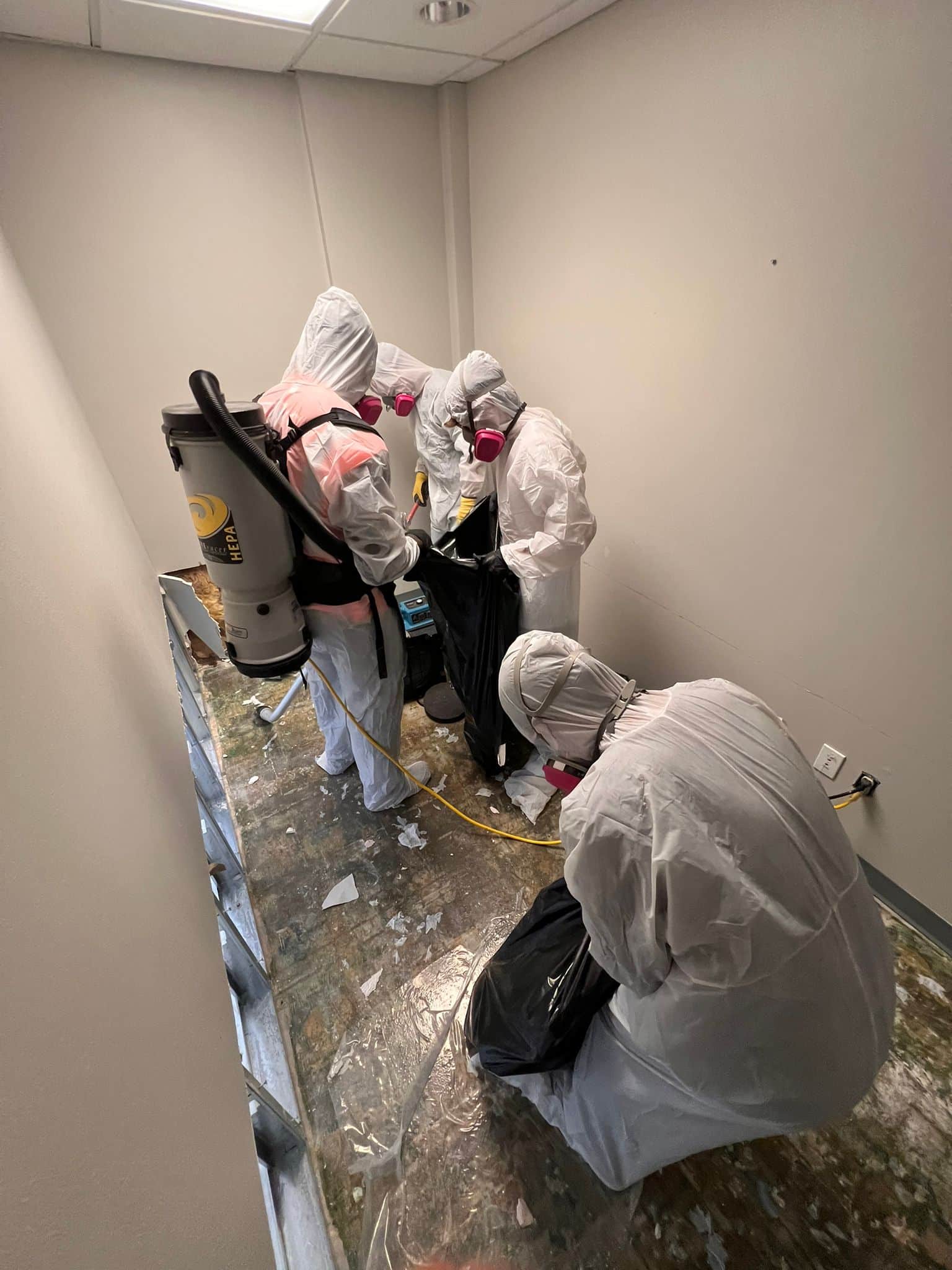 Three workers in white protective suits and pink respirator masks conduct remediation work in a confined indoor space. The floor is covered with plastic sheeting and debris, while equipment including a large cylindrical containment unit is visible on the left side. The minimal room with plain walls and fluorescent ceiling lighting suggests this is a hazardous materials or contamination cleanup operation.