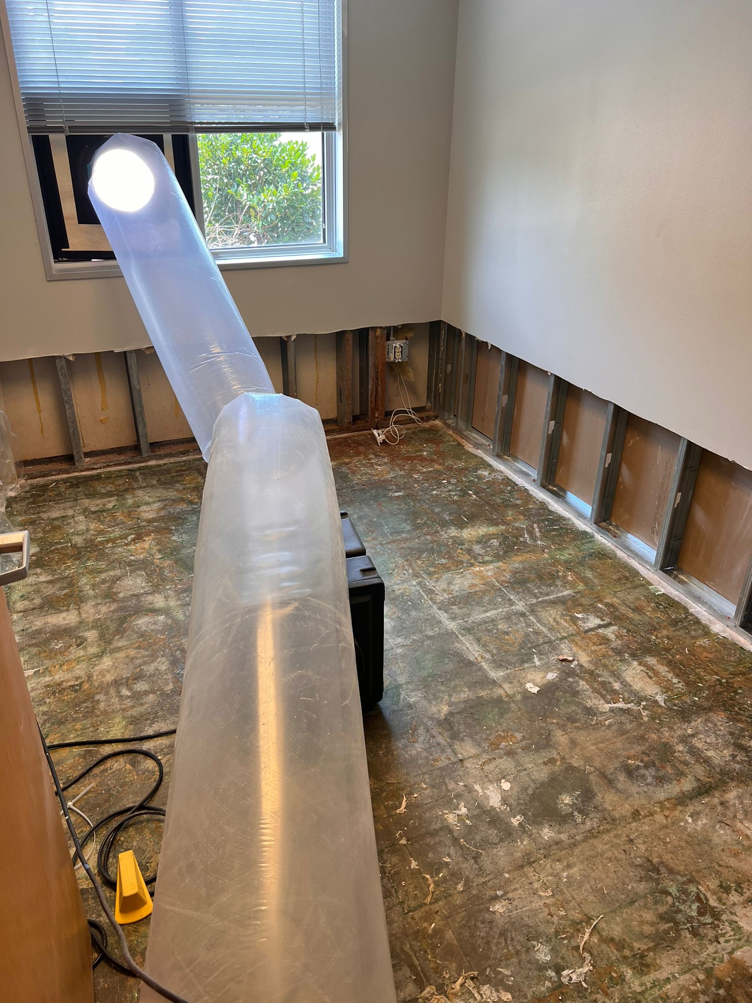 A clear plastic ventilation duct extends from a bright LED light source mounted on the window toward the basement floor of an unfinished room. The basement features exposed wooden framing, stained concrete flooring, and partially finished walls, with electrical outlets visible along the baseboards. The duct appears to be part of an air quality or dehumidification system being installed or tested in the residential space.