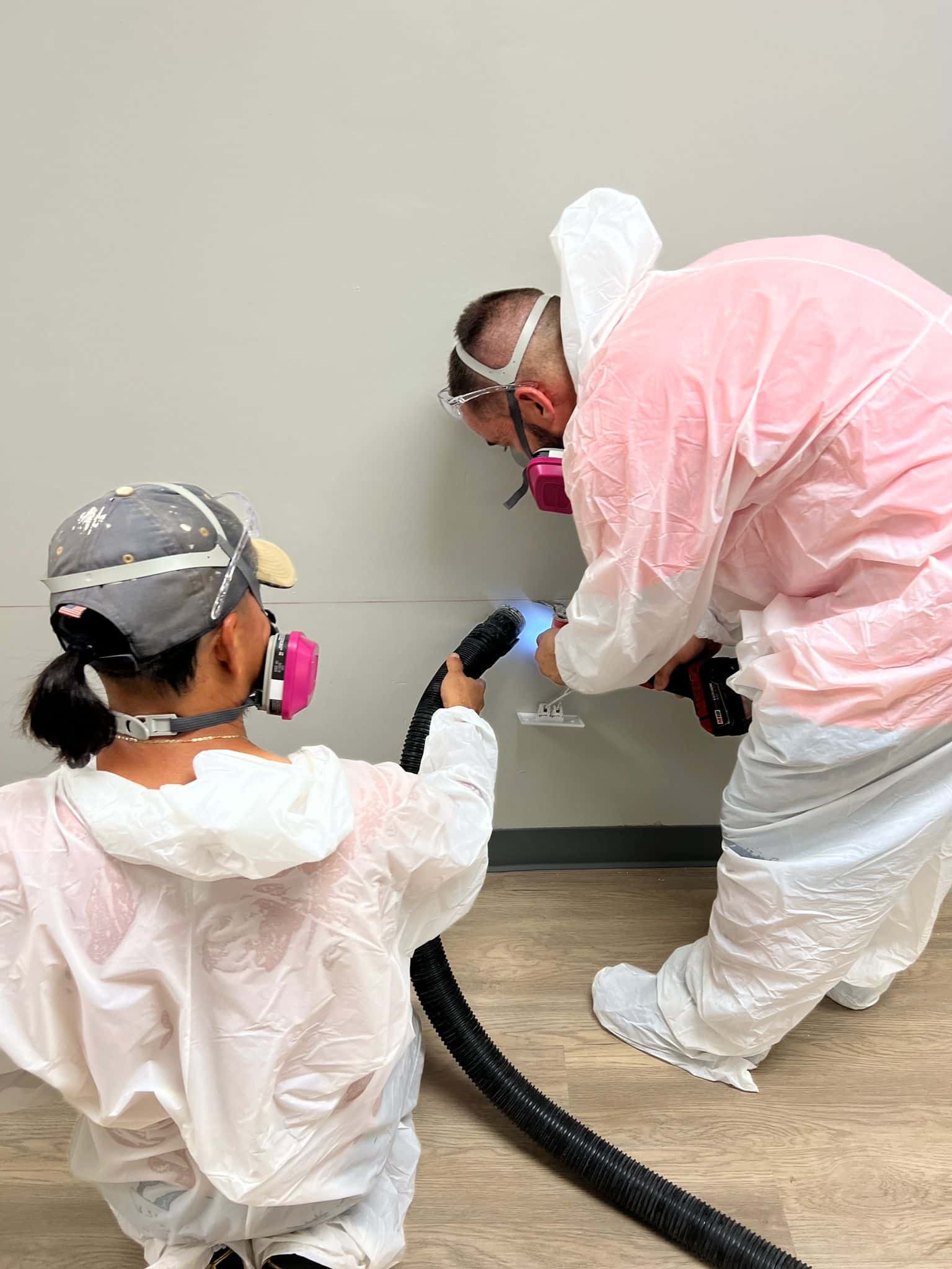 Two workers wearing protective equipment including white head coverings, respirators with pink filters, and white coveralls conduct what appears to be a spray painting or coating application task. One worker operates a spray gun while the other observes or assists in an indoor workspace with beige walls and wooden flooring. The protective gear and specialized equipment suggest this is an industrial or construction-related activity requiring safety precautions.