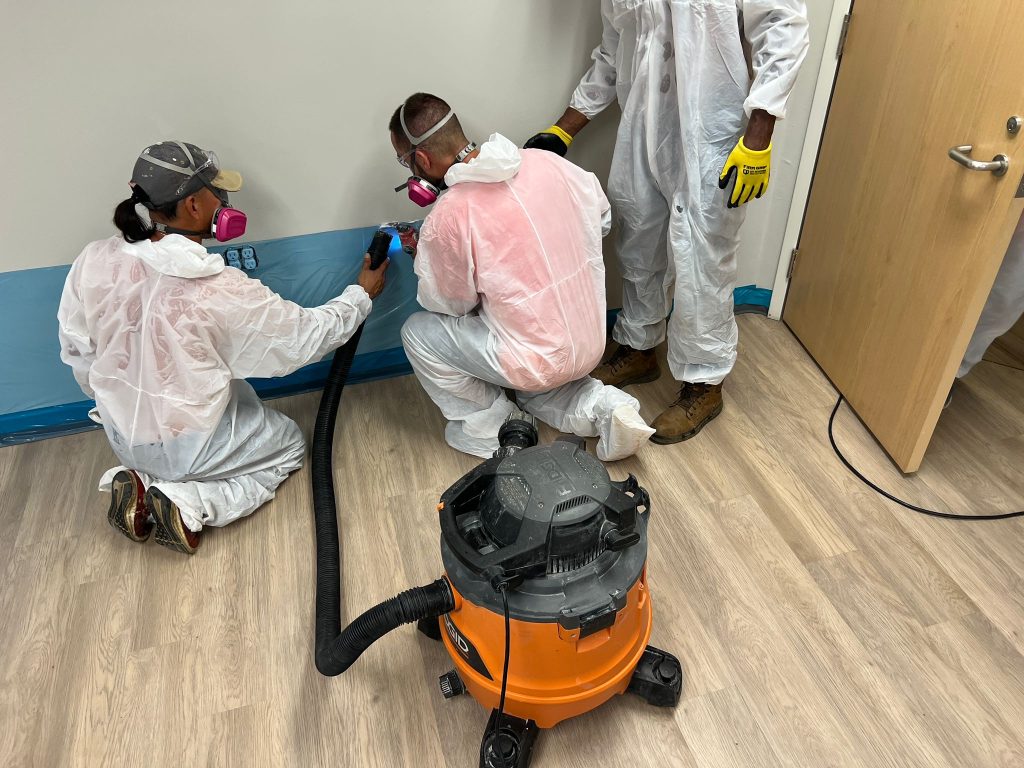 Three workers in full protective gear including white coveralls, respirators, and gloves use a wet vacuum system to conduct air quality or contamination testing in an indoor room. The orange and black wet/dry vacuum is positioned in the center with a hose extending across the space, while one worker operates testing equipment held in their hands. The scene appears to be part of a professional mold remediation, environmental assessment, or similar hazmat cleanup operation.