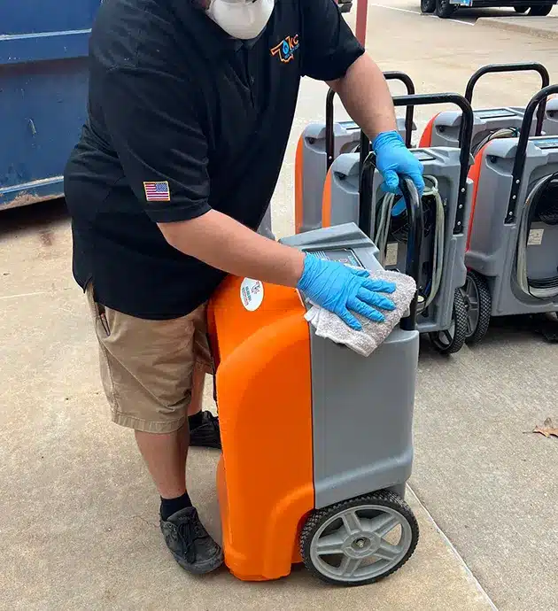 A person wearing a black shirt and blue gloves stands outdoors next to an orange and gray cart or cleaning equipment, appearing to be restocking or organizing supplies. The setting looks like a commercial or retail environment with vehicles visible in the background. The cart appears to be used for maintenance or cleaning services, suggesting this is a professional or facility management context.
