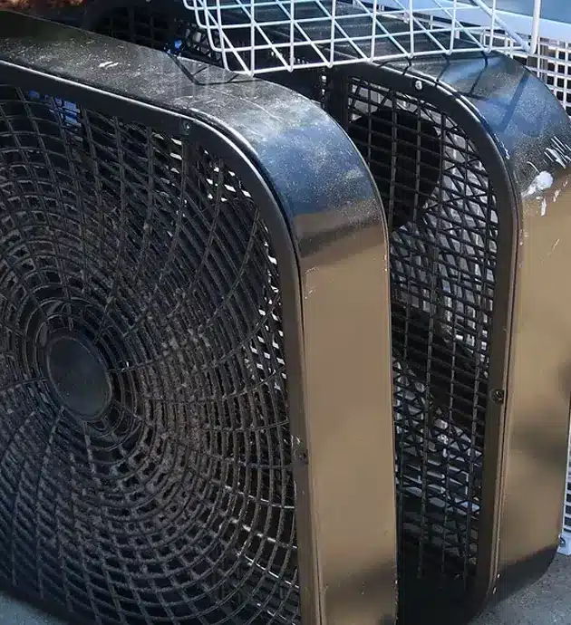 Two large industrial fan units with black mesh grilles and metallic silver casings are positioned side by side, likely in a storage or warehouse setting. The fans feature prominent circular mesh screens on their front faces and appear to be heavy-duty cooling or ventilation equipment. A wire storage rack is partially visible in the background above the units.