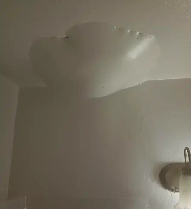 A bathroom interior with white walls and ceiling featuring visible water stains and discoloration, indicating moisture damage or mold growth. A round exhaust vent or fixture is visible on the right side of the wall. The poor lighting and hazy appearance suggest humidity or ventilation issues within the space.