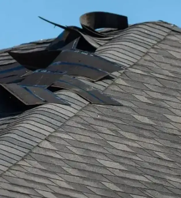 A residential roof with asphalt shingles shows significant storm damage, with multiple shingles torn away and displaced, exposing the underlying roof structure. A black roof vent or pipe protrudes from the damaged area against a clear blue sky. The damage appears to be from severe wind or weather impact, highlighting the need for roof repair or replacement.