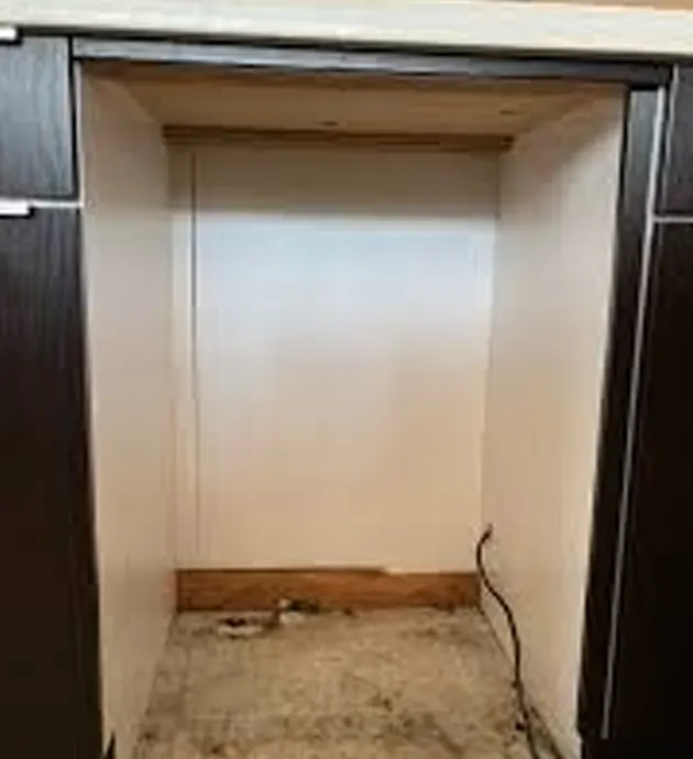 A narrow, empty closet or storage space with cream-colored walls and a concrete floor, framed by dark wood or metal trim on the sides. The interior appears bare except for a single electrical outlet or wire visible on the right wall. The space looks unfinished or awaiting renovation.