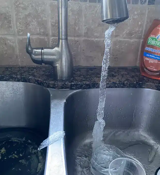 A kitchen sink with two stainless steel basins is shown with water running from the faucet into the right basin. The sink appears to be in use with water actively flowing, and cleaning supplies are visible on the counter behind it. The backsplash features a tiled pattern typical of kitchen installations.