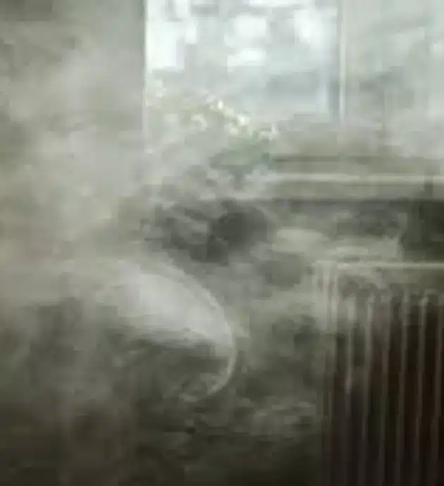 A heavily obscured interior room filled with thick smoke or steam, with a window visible on the right side and a radiator beneath it. The smoke is so dense that details are barely discernible, creating a hazy, almost surreal atmosphere. The image appears to capture a moment of extreme fog or smoke accumulation in what looks like a residential or institutional space.