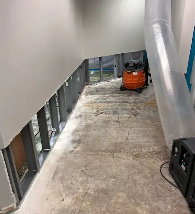 A narrow basement or utility corridor with concrete flooring, white walls, and a metal handrail running along one side. The space contains various items including storage boxes, an orange equipment case, and what appears to be construction or maintenance materials scattered throughout. A white PVC pipe runs along the ceiling, suggesting active plumbing or HVAC work in progress.