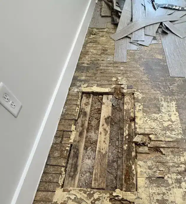 A flooded interior room with water damage showing exposed wooden floorboards and structural elements beneath damaged flooring. The white drywall on the left wall contrasts with the wet, deteriorated wood subfloor in the center. Electrical wiring and building materials are visible along the ceiling, indicating ongoing water damage assessment or restoration work.
