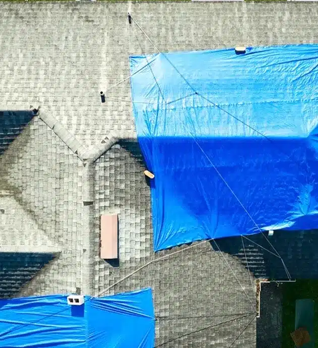 An aerial view of a building exterior under renovation or weatherproofing, with large sheets of blue plastic sheeting covering portions of the brick facade and roof. The protective covering is secured with ropes and appears to span multiple sections of the structure, suggesting ongoing construction or repair work. A small pink door or window frame is visible on the brick wall beneath the plastic covering.