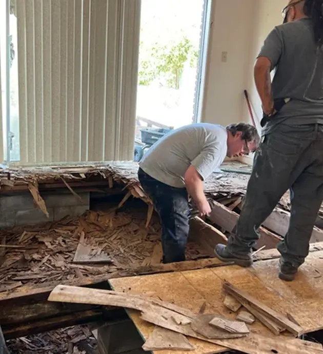 Two workers examine severe water damage and deteriorating wooden flooring in an indoor residential space. The man in the center bends down to inspect the warped and splintered wood debris scattered across the floor, while another worker in dark clothing stands nearby. Vertical blinds and natural light from windows are visible in the background of what appears to be a home restoration or damage assessment project.