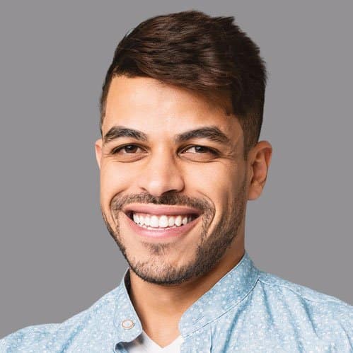 A smiling man with dark brown hair and facial hair poses for a professional headshot against a neutral gray background. He wears a light blue button-up shirt and displays a bright, genuine smile. The image appears to be a standard portrait photo suitable for professional or business purposes.