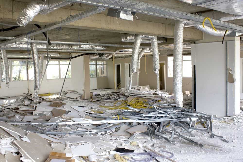 An interior commercial or industrial space undergoing demolition or renovation, with debris scattered across the floor including drywall, metal ducts, and electrical wiring. The open ceiling exposes structural concrete beams and support columns wrapped in insulation, while large windows line the walls allowing natural light to flood the vacant area. Yellow safety markings and caution tape are visible among the construction waste.