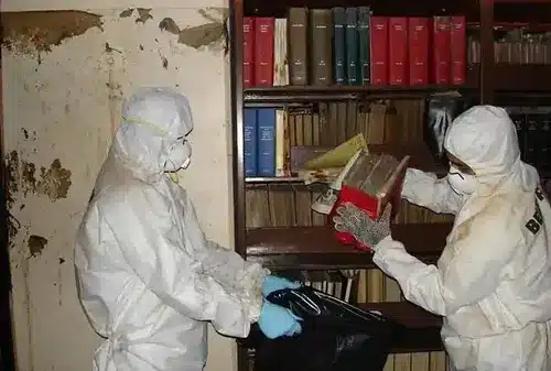 Two people in full protective suits, including white coveralls and blue gloves, work inside a library or study room surrounded by bookshelves containing leather-bound volumes and reference materials. They appear to be conducting some kind of decontamination, remediation, or hazardous material handling procedure, with one person holding what looks like collection equipment or documentation. The aged wall behind them shows significant water damage and discoloration, suggesting the work may be related to restoration or contamination cleanup.
