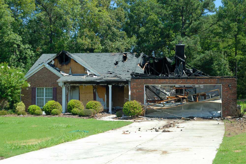 A severely damaged brick house with a collapsed roof and charred remains, showing extensive fire damage to the structure and garage. The front entrance is boarded up, and the garage door opening exposes burnt debris and metal framework. The property is surrounded by well-maintained landscaping with manicured shrubs and green lawn, creating a stark contrast to the destruction of the home.