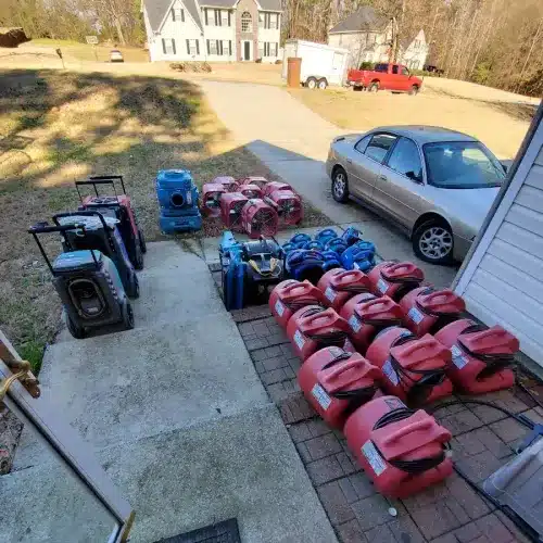 Aerial view of a residential driveway displaying numerous red and blue storage containers or equipment cases arranged in rows on a brick patio. A silver SUV is parked nearby, with additional vehicles visible in the background along with a two-story white house and bare trees, suggesting a spring or early season timeframe.