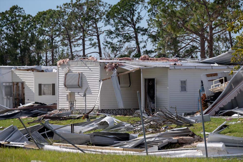 A mobile home severely damaged by what appears to be a hurricane or tornado, with the roof partially torn off, windows blown out, and debris scattered across the surrounding grass and yard. The white single-wide trailer sits among pine trees with visible structural damage to its walls and exterior fixtures. Debris including metal roofing, siding, and household materials are strewn across the ground in front of the destroyed residence.