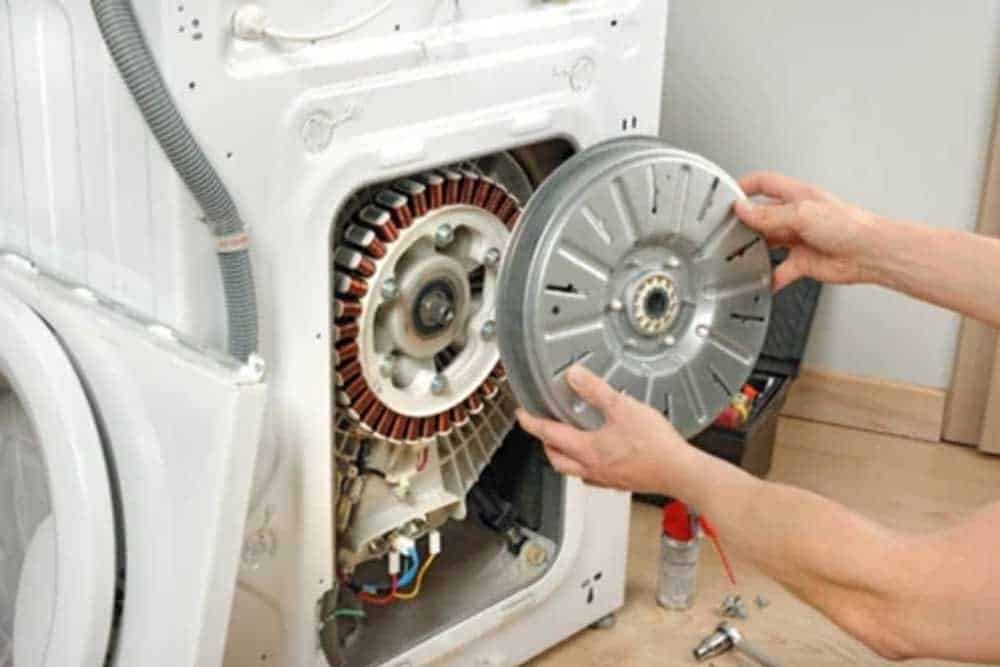 A person's hands remove the gray drum from the interior of a white clothes dryer, revealing the red heating element and internal drum mechanism. The dryer is disassembled for maintenance or repair, with various components visible including the motor and wiring. A spray bottle and screwdriver are visible on the floor beside the appliance, indicating active servicing work.