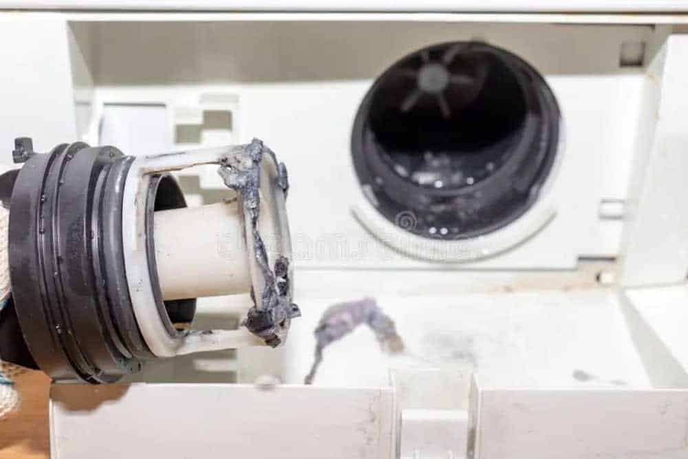 A disassembled washing machine drum seal and gasket lies on an open washing machine, showing accumulated lint and debris around the rubber components. The drum's interior is visible in the background, displaying the metallic drum surface. This image depicts routine maintenance or repair work being performed on a front-loading washer.