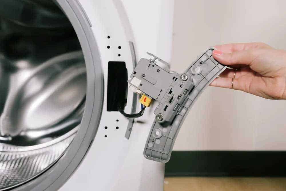 A person's hand holding the inner door latch assembly of a washing machine, showing the mechanical components including a yellow connector and metal parts. The disassembled piece is being removed from the white washing machine door, demonstrating appliance maintenance or repair work.