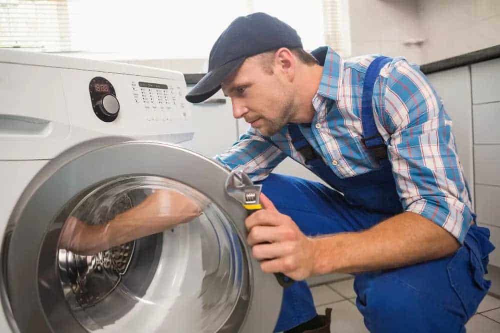 A male repair technician wearing a blue plaid shirt, blue overalls, and dark cap inspects the interior drum of a front-load washing machine while holding a flashlight. He is crouched down in a residential laundry room, examining the appliance's components during what appears to be a maintenance or repair service call.