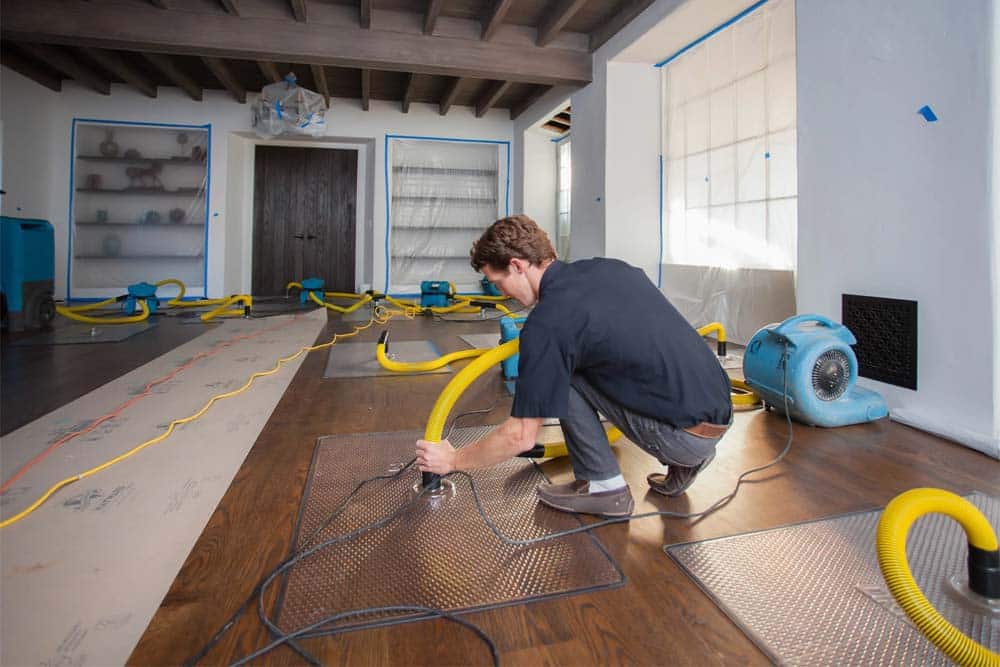 A worker in a dark navy shirt crouches on a wooden floor while operating a power tool to install or repair yellow and black ducting hoses laid out in a pattern across the room. The interior space features exposed beam ceilings, white walls with blue trim, and multiple doorways, with a blue air circulation fan visible on the right side. The setup appears to be part of a water damage restoration or HVAC installation project, with protective plastic sheeting covering portions of the floor and walls.