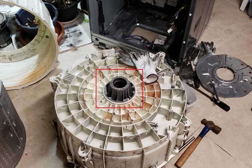 A disassembled washing machine drum sits on a workshop floor during repair, with its large cream-colored outer tub displayed upside down and a dark inner drum highlighted by a red rectangle in the center. Various components including the rear panel, gaskets, and tools are scattered around the workspace, indicating mid-repair maintenance work. The setting appears to be an appliance repair shop with additional equipment and machinery visible in the background.