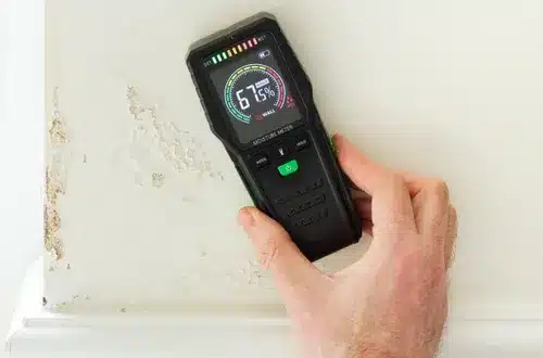 A person holds a digital moisture meter against a white wall showing visible signs of water damage and peeling paint. The handheld device displays a moisture reading of 95.5% on its illuminated screen, indicating high moisture content in the wall material. The meter has a black casing with various buttons and a digital display showing measurement data and a circular gauge.