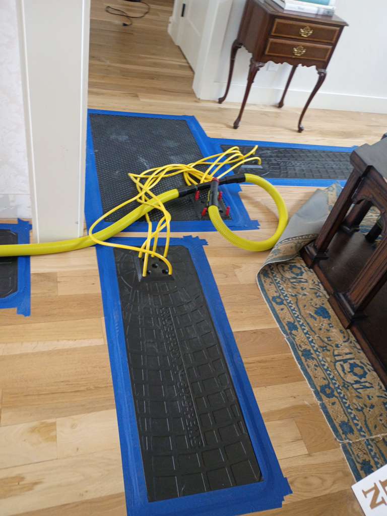 A yellow extension cord is coiled on top of a blue-bordered black anti-fatigue mat laid across a wooden floor in an interior space. The setup appears to be part of a water damage restoration or drying operation, with additional blue protective materials visible in the background. The room contains period furniture including a wooden side table with brass hardware and a decorative runner rug along the baseboard.
